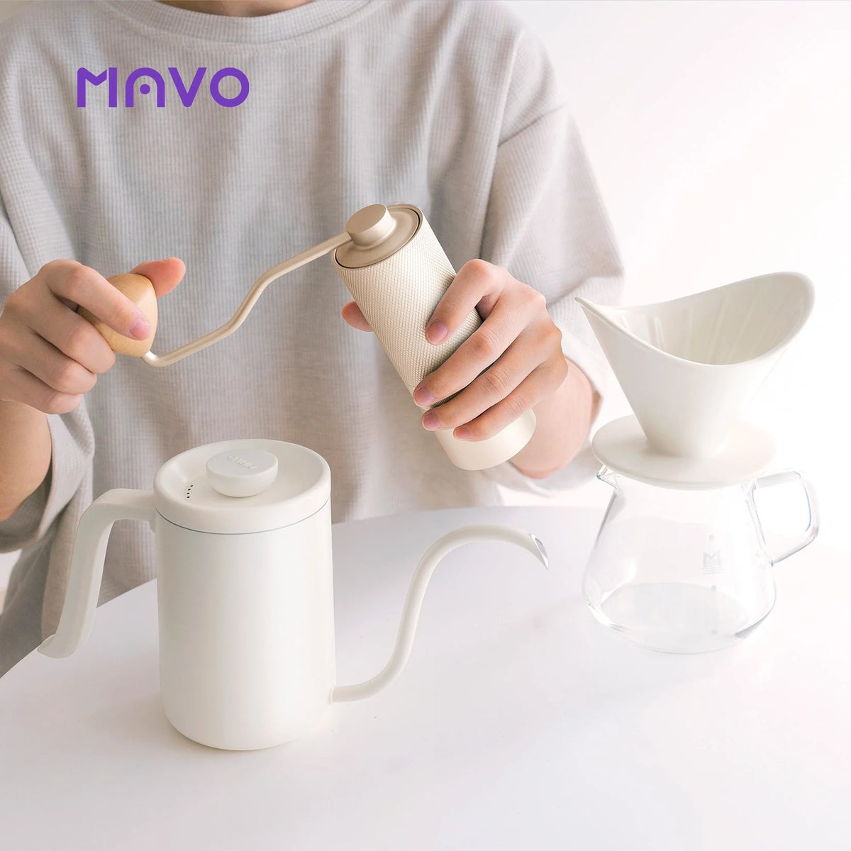 Grinding beans with the silver Wizard hand grinder next to the light-coloured Deep Flow Pro kettle and dripper on a glass server