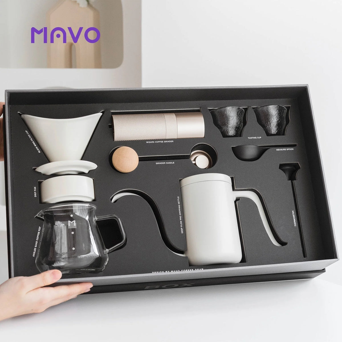 Hands holding the open MAVO gift box with the light-coloured set fitted in foam: grinder, kettle, dripper, glass server, cups, spoon and thermometer