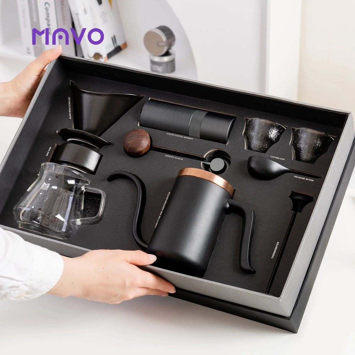 Hands holding the open MAVO gift box with the dark set fitted in foam: grinder, copper-rimmed kettle, dripper, glass server, cups, spoon and thermometer