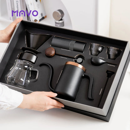 Hands holding the open MAVO gift box with the dark set fitted in foam: grinder, copper-rimmed kettle, dripper, glass server, cups, spoon and thermometer