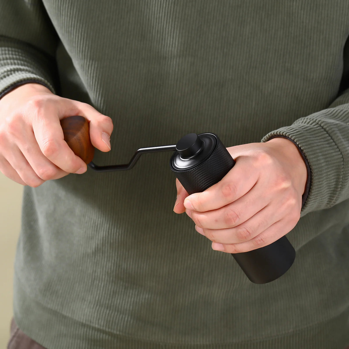 Hands turning the crank of the black MAVO Wizard 2.0 hand grinder, dark wood knob on the S-shaped S-shaped handle