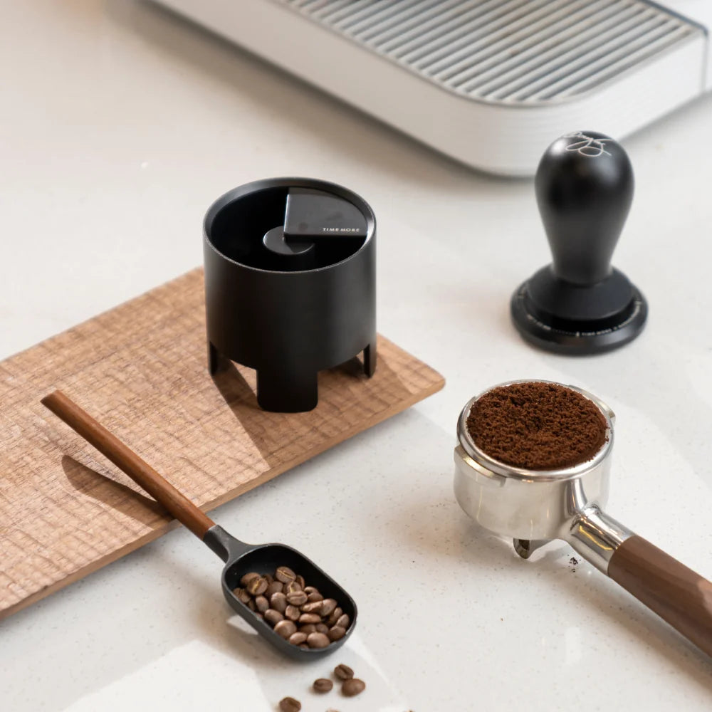 The black Autospin WDT Tool standing on a wooden board beside a black tamper, a portafilter of ground coffee and a bean scoop