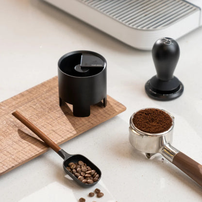 The black Autospin WDT Tool standing on a wooden board beside a black tamper, a portafilter of ground coffee and a bean scoop