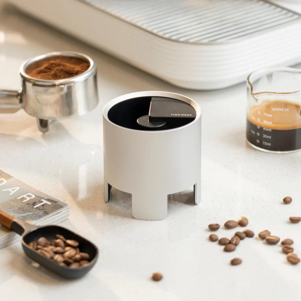 The silver Autospin WDT Tool standing on a counter in front of a portafilter of ground coffee and a glass of espresso, with scattered beans