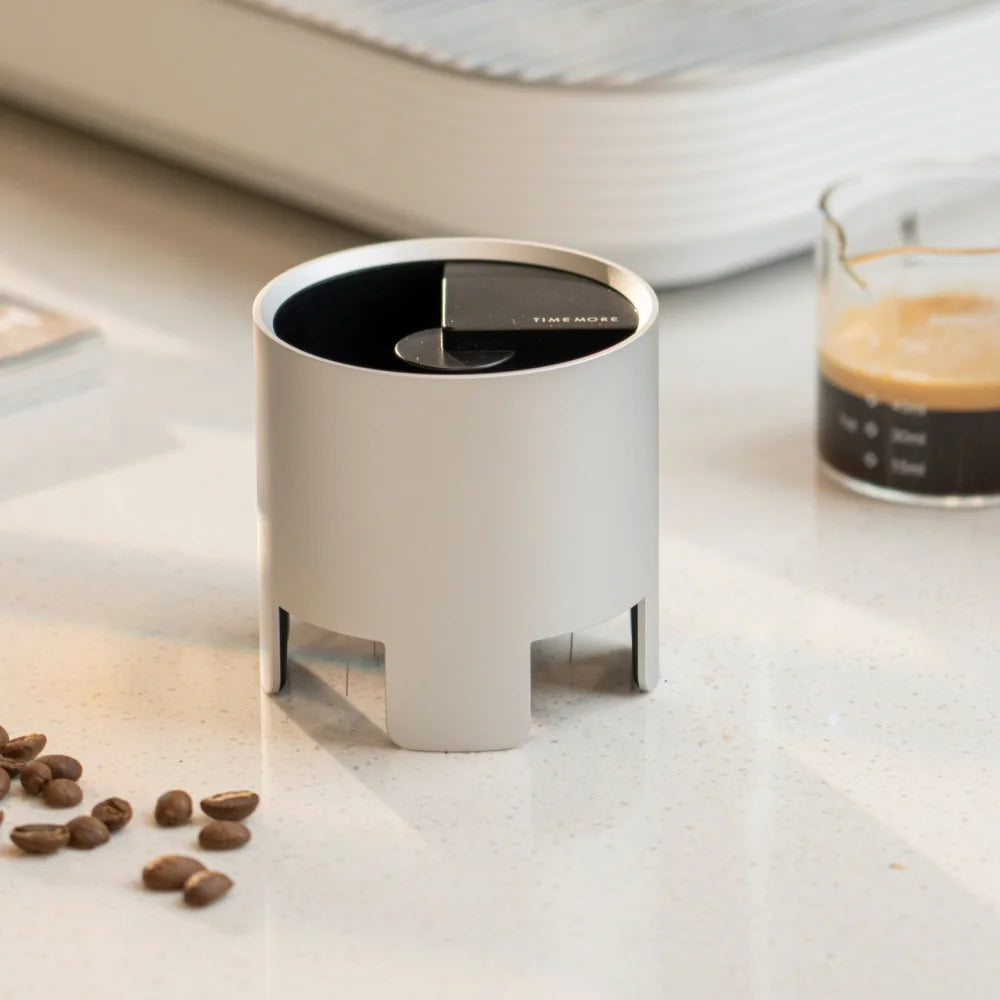 The silver Autospin WDT Tool standing on a white counter beside a glass of espresso and coffee beans