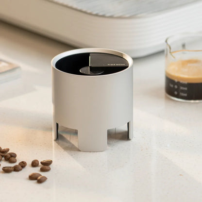 The silver Autospin WDT Tool standing on a white counter beside a glass of espresso and coffee beans