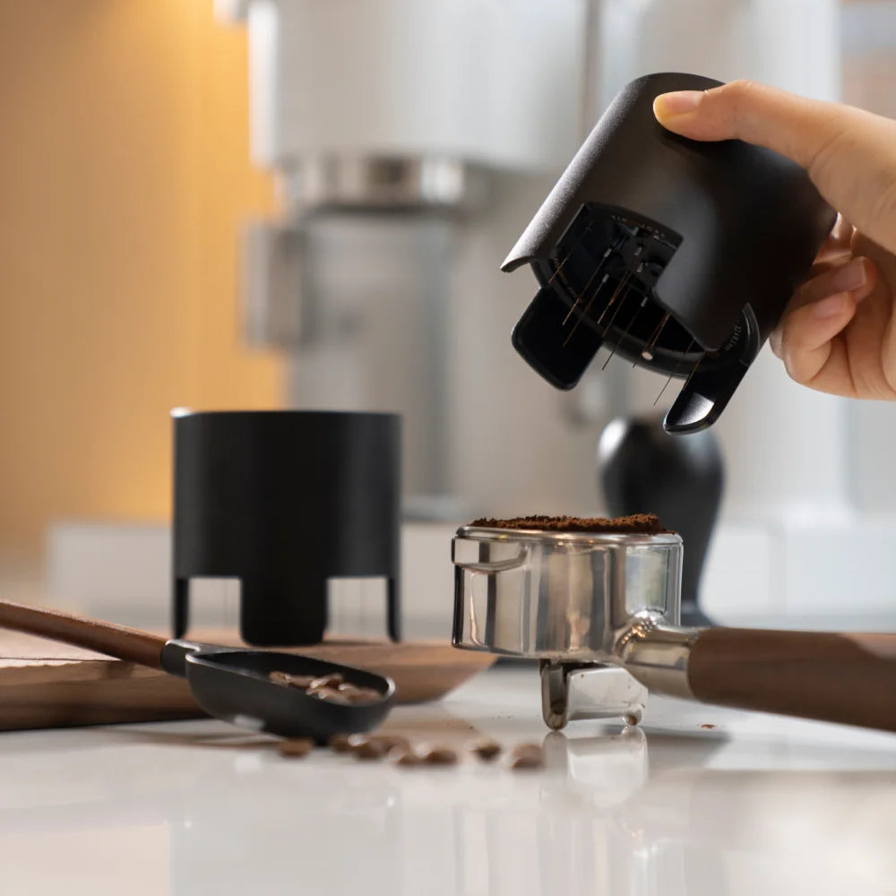 A hand lifting the black Autospin WDT Tool away from a portafilter of ground coffee on a white counter, with a bean scoop and a second Autospin behind it