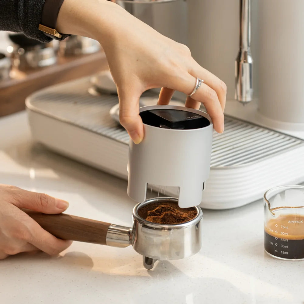 A hand holding the silver Autospin WDT Tool in place on a portafilter in front of an espresso machine, with a shot glass of espresso beside it