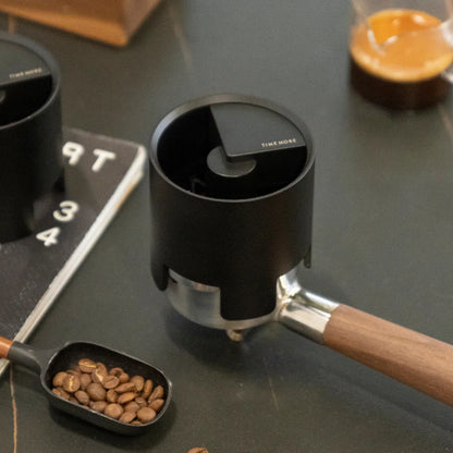 The black Autospin WDT Tool resting on a portafilter beside a scoop of coffee beans on a dark marble counter