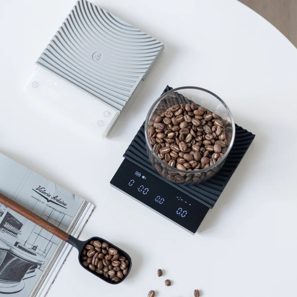 Flat lay of the white and black TIMEMORE Basic 3 scales with a glass jar of coffee beans, a bean scoop and a magazine