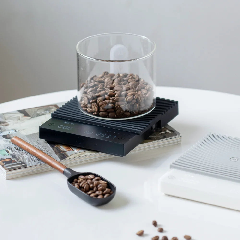 A glass jar of coffee beans being weighed on the black TIMEMORE Basic 3, which sits on a stack of magazines beside the white version