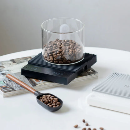 A glass jar of coffee beans being weighed on the black TIMEMORE Basic 3, which sits on a stack of magazines beside the white version