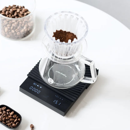 Pour-over brewing on the black TIMEMORE Basic 3: a glass dripper of ground coffee sits on a glass carafe on the scale, with the coffee-to-water ratio shown as 15.1 on the display