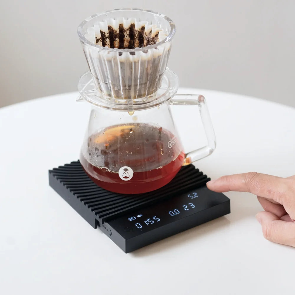 Coffee dripping into a glass carafe on the black TIMEMORE Basic 3 in pour-over mode, a finger reaching for the touch controls as time and weight run on the display
