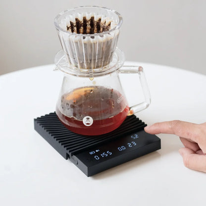 Coffee dripping into a glass carafe on the black TIMEMORE Basic 3 in pour-over mode, a finger reaching for the touch controls as time and weight run on the display