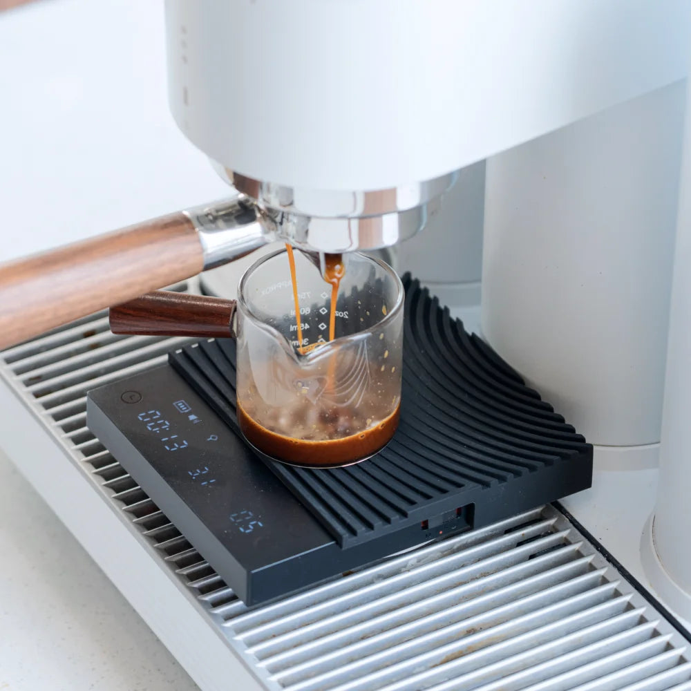 The black TIMEMORE Basic 3 on an espresso machine drip tray with the mat flipped to its contoured portafilter rest, weighing a shot pulled into a glass cup