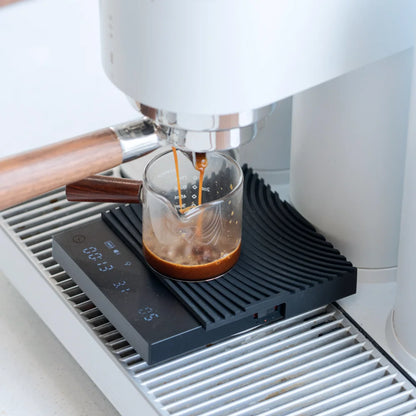 The black TIMEMORE Basic 3 on an espresso machine drip tray with the mat flipped to its contoured portafilter rest, weighing a shot pulled into a glass cup