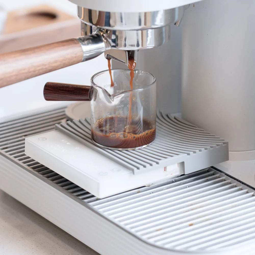 The white TIMEMORE Basic 3 under an espresso machine group head, catching a shot in a glass cup on its white silicone mat