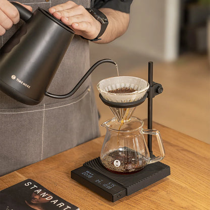 A hand pours from a black TIMEMORE gooseneck kettle into a dripper held in the DUO's ring above a glass server on the scale, on a wooden table