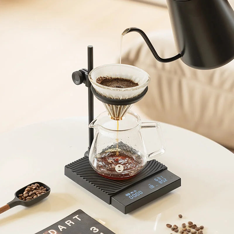Pour-over in progress on the DUO: a kettle pouring into a glass dripper in the swivel ring above a glass server, with coffee beans and a wooden scoop beside the scale