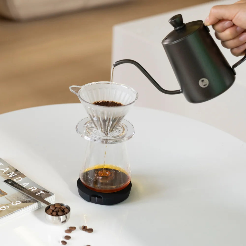 Black TIMEMORE Black Mirror Dot weighing platform under a glass server on a white table, a pour-over dripper of ground coffee on top, a black gooseneck kettle pouring and a bean scoop beside it