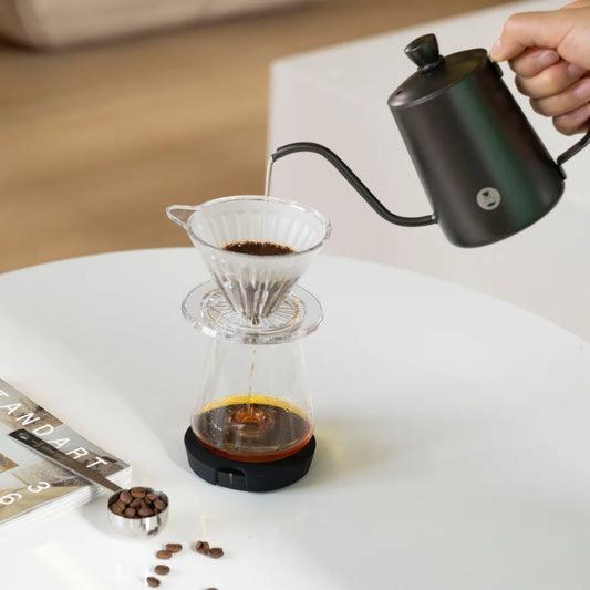 Black TIMEMORE Black Mirror Dot weighing platform under a glass server on a white table, a pour-over dripper of ground coffee on top, a black gooseneck kettle pouring and a bean scoop beside it