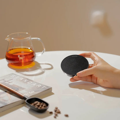 A hand holding a black TIMEMORE Dot disc in front of a glass server of coffee, a bean scoop and a magazine
