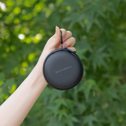 A hand holding the TIMEMORE Black Mirror Dot in its round black zip travel case outdoors in front of green foliage