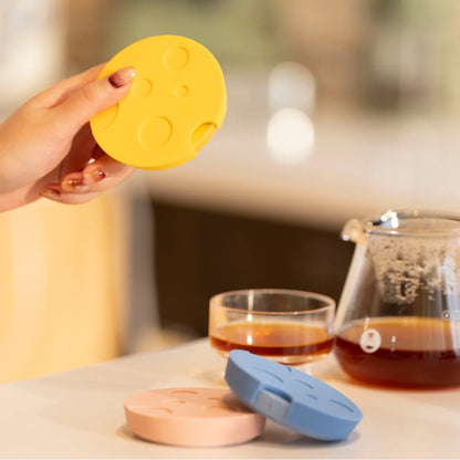 A hand holding a yellow TIMEMORE Dot disc with a dimpled crater-pattern silicone mat, pink and blue Dots and glass servers of coffee behind it