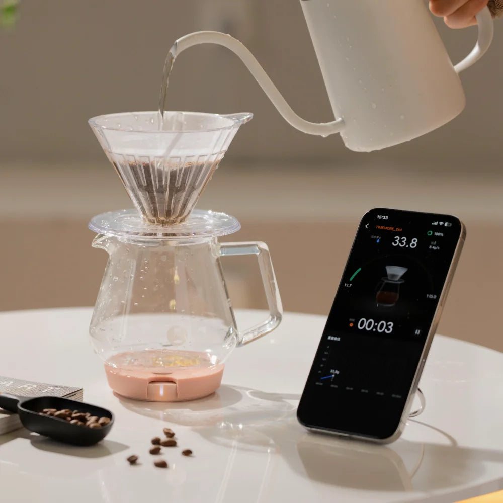 Water pouring from a white kettle into a pour-over dripper on a glass server standing on a pink Dot, with a phone beside it showing the TIMEMORE app timer at 00:03 and 33.8g