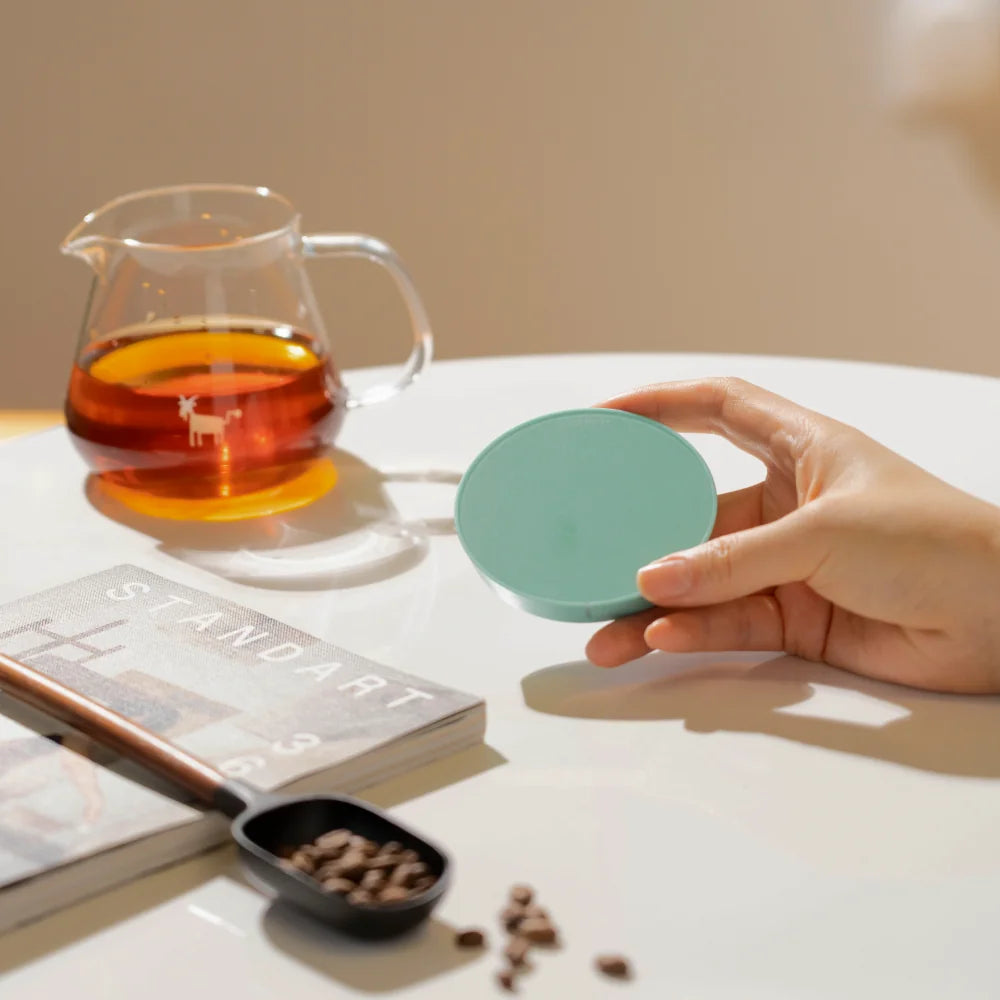 A hand holding a green TIMEMORE Dot disc beside a glass server of coffee and a magazine on a white table