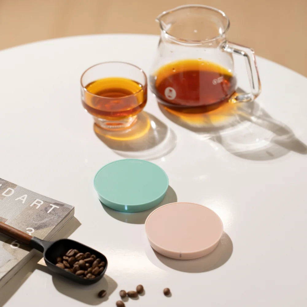 Green and pink TIMEMORE Dot discs lying on a white table beside a glass of coffee, a glass server and a scoop of coffee beans
