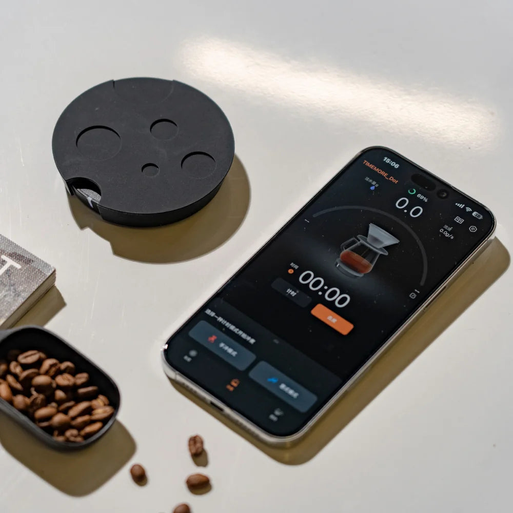 The black TIMEMORE Dot with its crater-pattern silicone mat next to a phone running the TIMEMORE app at 00:00 and 0.0g, with a scoop of coffee beans on the table