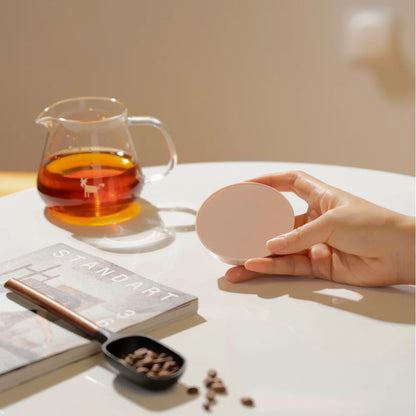 A hand holding a pink TIMEMORE Dot disc in front of a glass server of coffee and a magazine