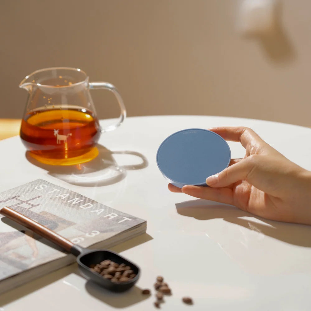 A hand holding a blue TIMEMORE Dot disc in front of a glass server of coffee and a magazine