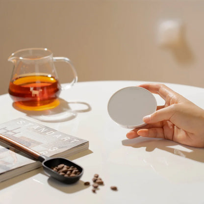 A hand holding a white TIMEMORE Dot disc in front of a glass server of coffee and a magazine