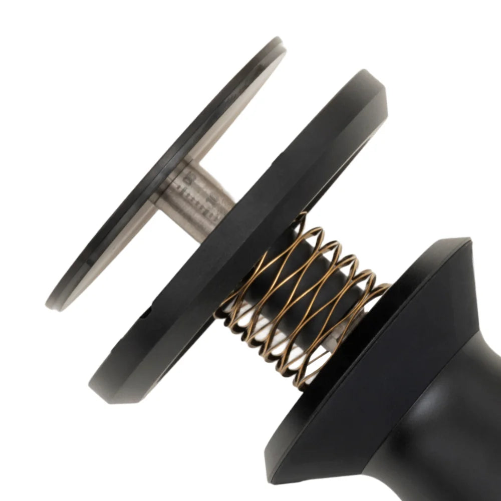 Close-up of the FLOW tamper's gold wave spring between the black base and collar, shown at an angle against a white background