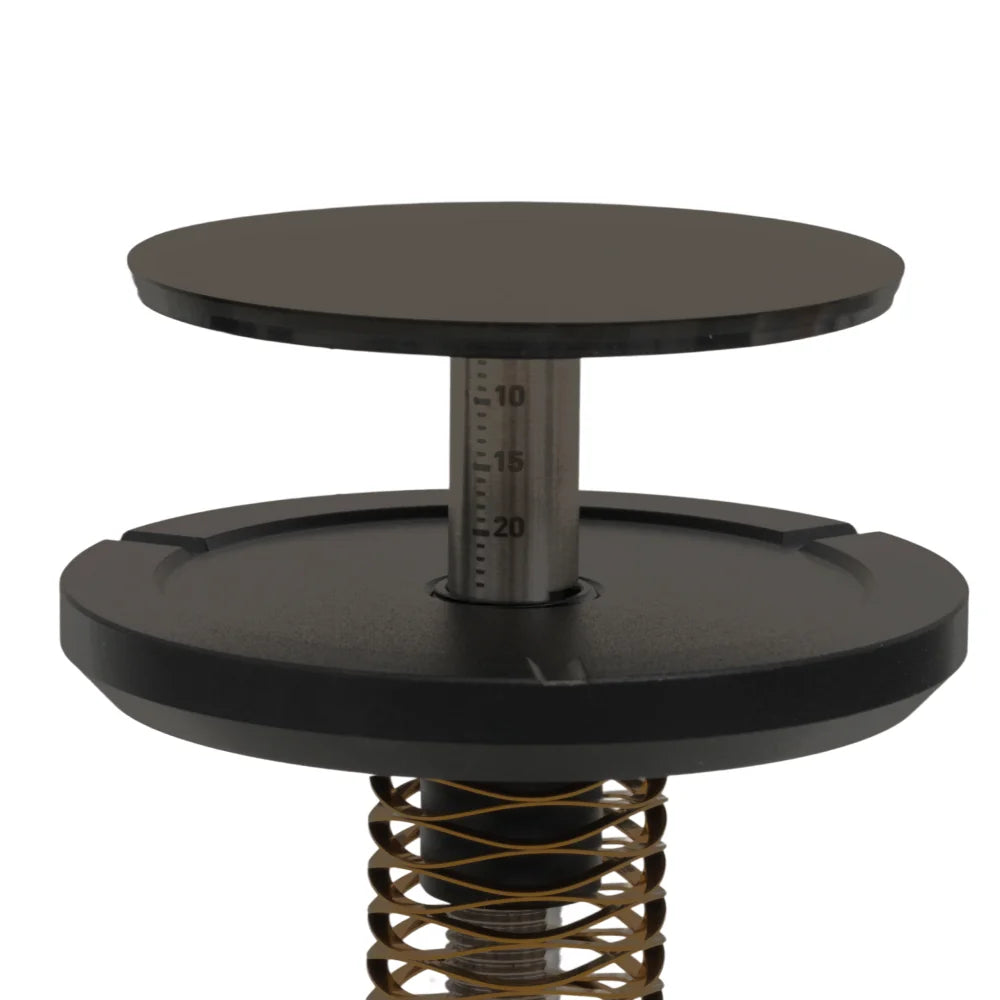 Close-up of the FLOW tamper's depth dial: a stainless shaft with a millimetre scale rising from the gold wave spring to the black base plate