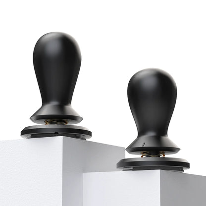 Two FLOW tampers standing on white plinths, each with a gold wave spring visible between the collar and the base