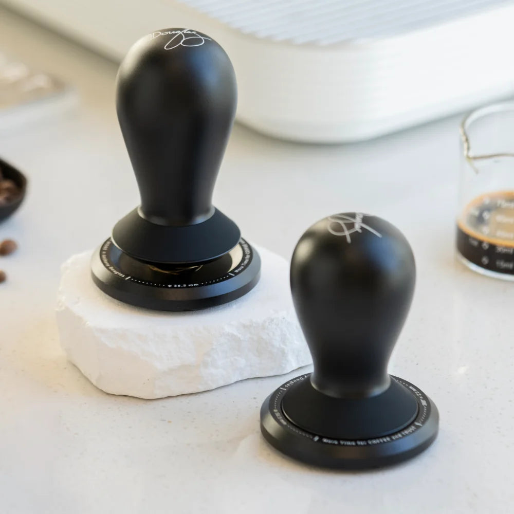 Two black FLOW tampers on a white stone surface beside an espresso glass, the handles signed by Anthony Douglas
