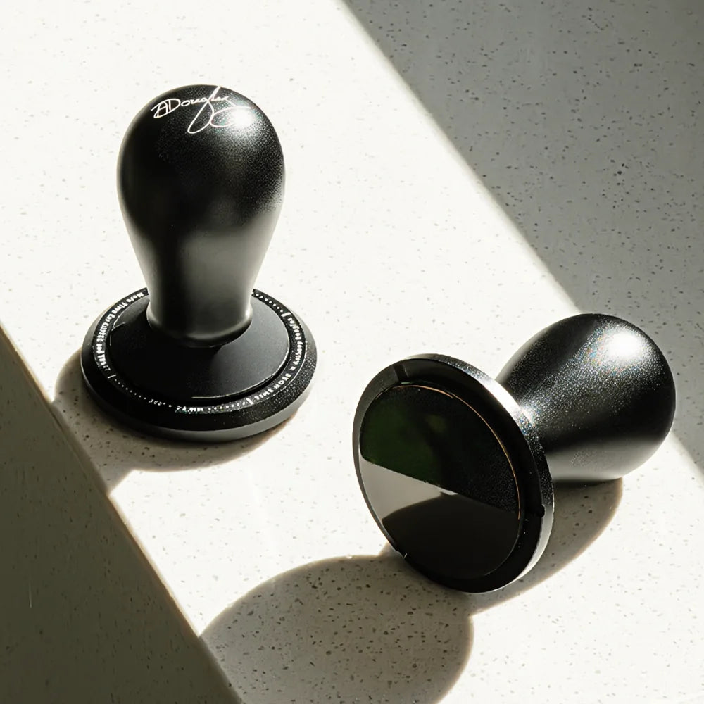 One FLOW tamper standing in sunlight and a second lying on its side to show its mirror-black DLC base on a white countertop
