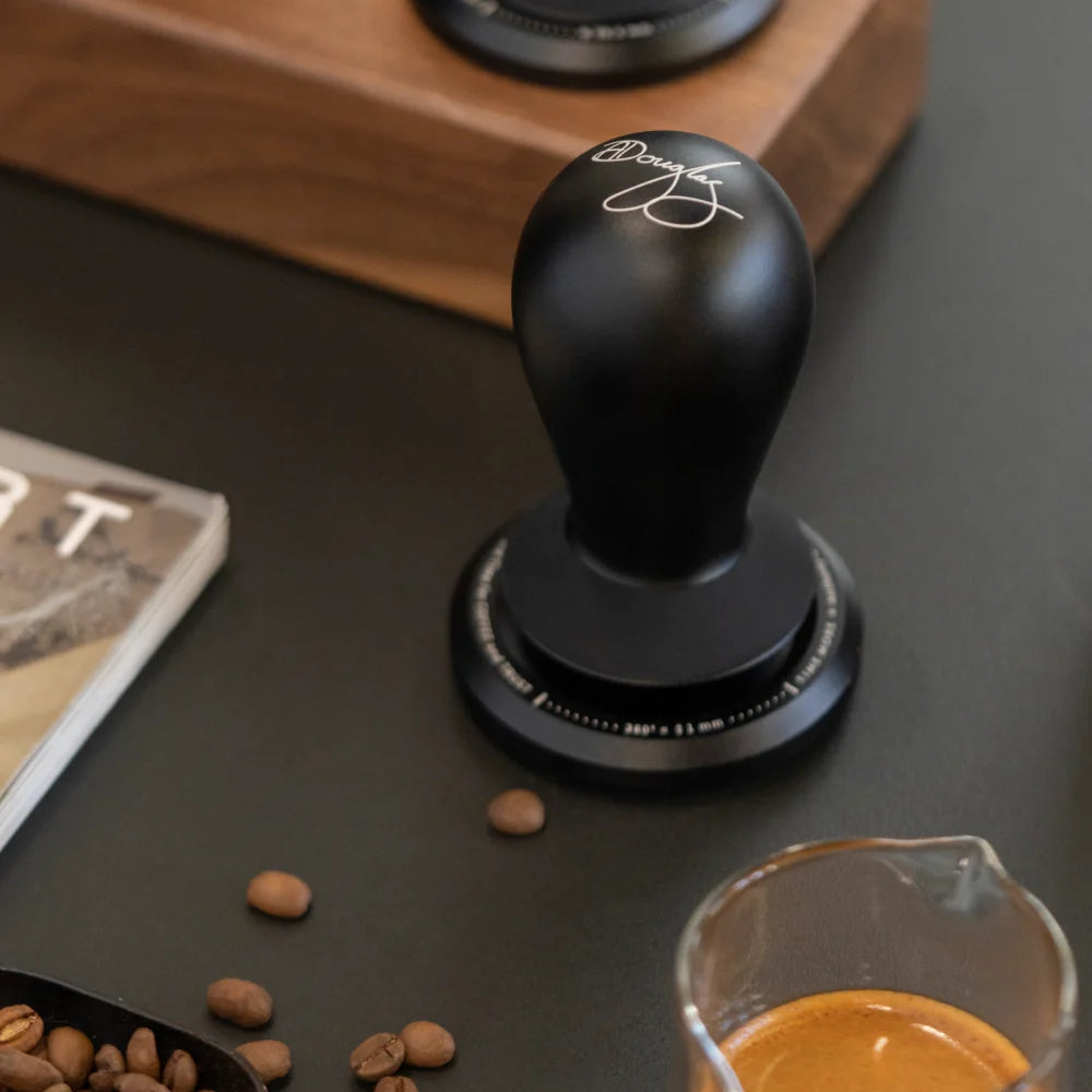 A FLOW tamper standing on a dark counter beside scattered coffee beans, a scoop and a glass of espresso