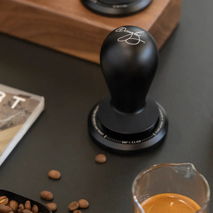 A FLOW tamper standing on a dark counter beside scattered coffee beans, a scoop and a glass of espresso