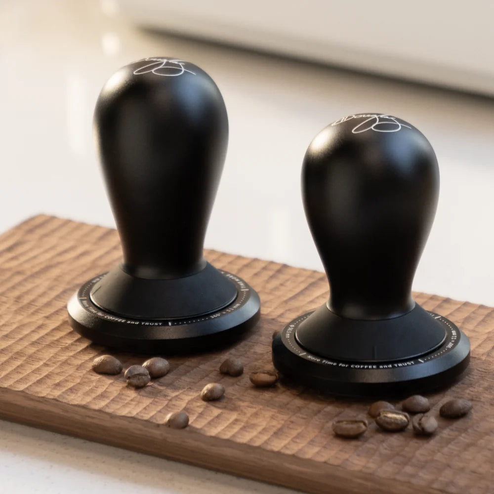 Two FLOW tampers side by side on a carved wooden board with coffee beans scattered around them