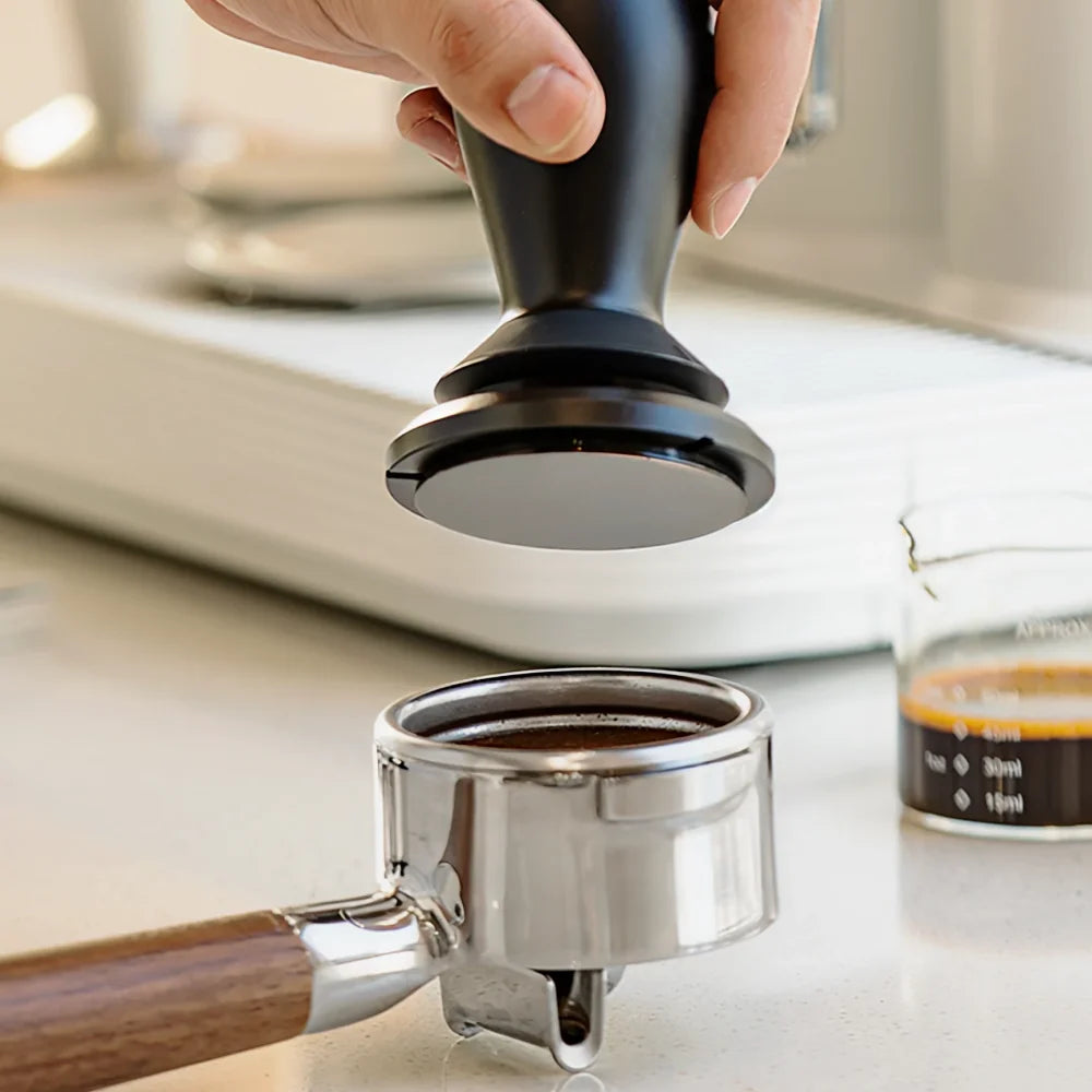 A hand holding the FLOW tamper above a stainless steel portafilter filled with ground coffee on a white counter