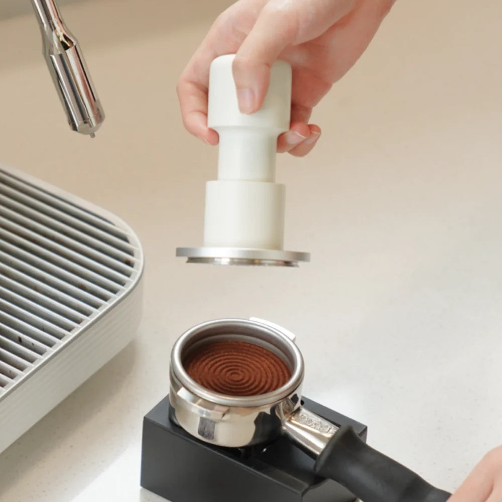A hand holding the white Impulse Impact Tamper above a portafilter of ground coffee, with an espresso machine behind