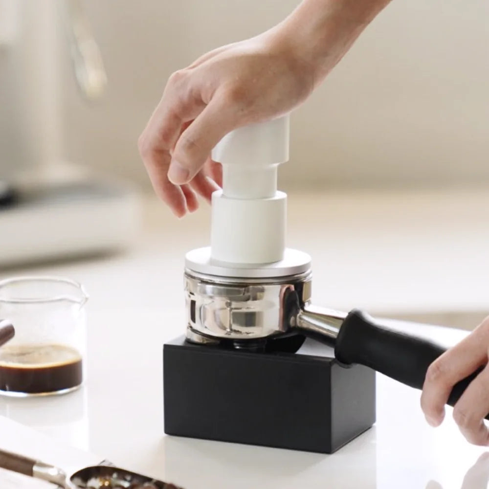A hand pressing the white Impulse Impact Tamper into a portafilter held in a black tamping stand, with a shot glass of espresso beside it