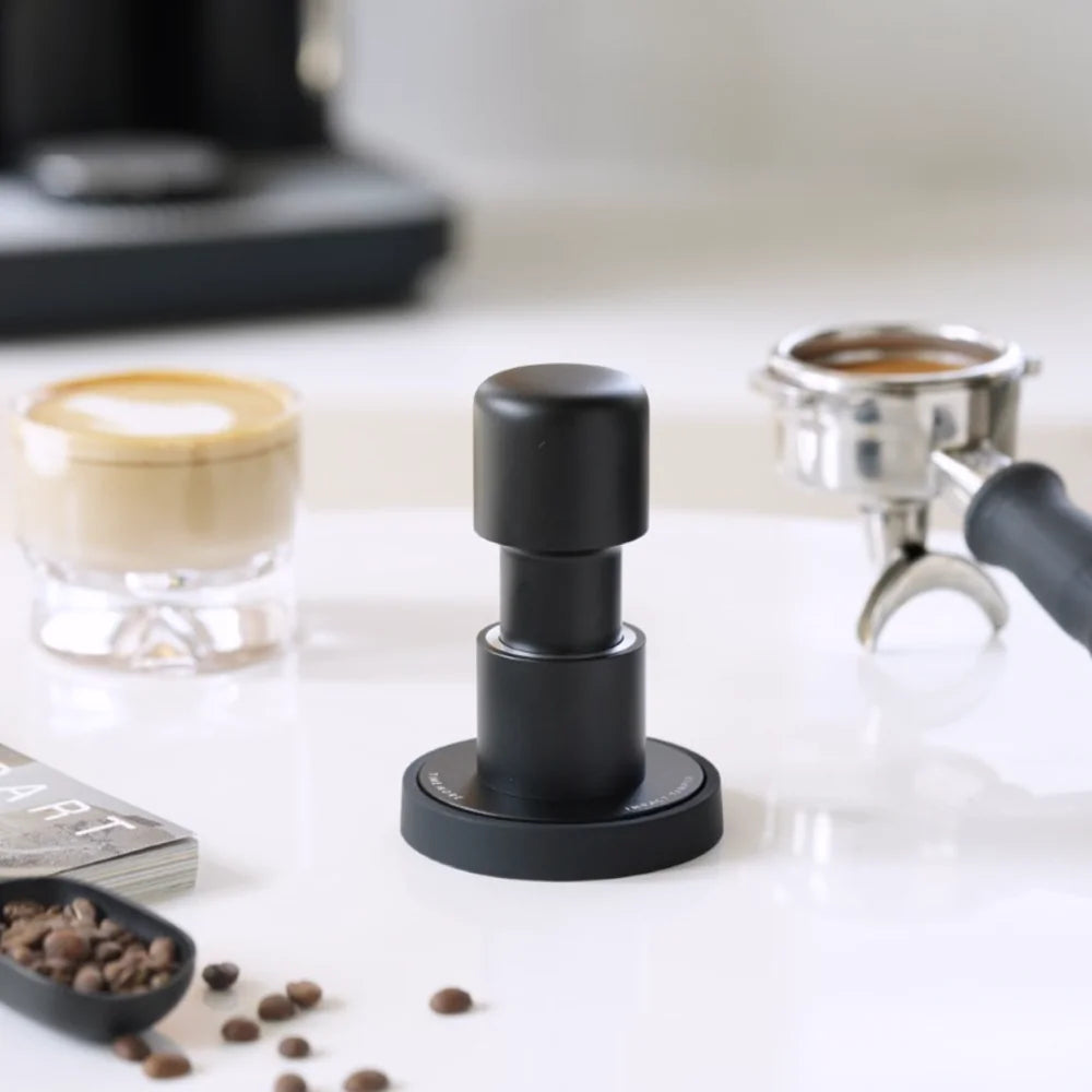 Black Impulse Impact Tamper standing on a white counter beside a portafilter, a latte and a scoop of coffee beans
