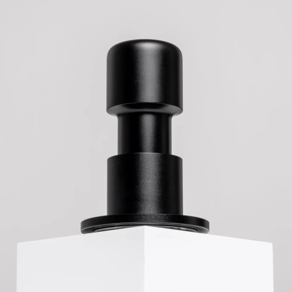 Black Impulse Impact Tamper standing on a white plinth against a light grey wall