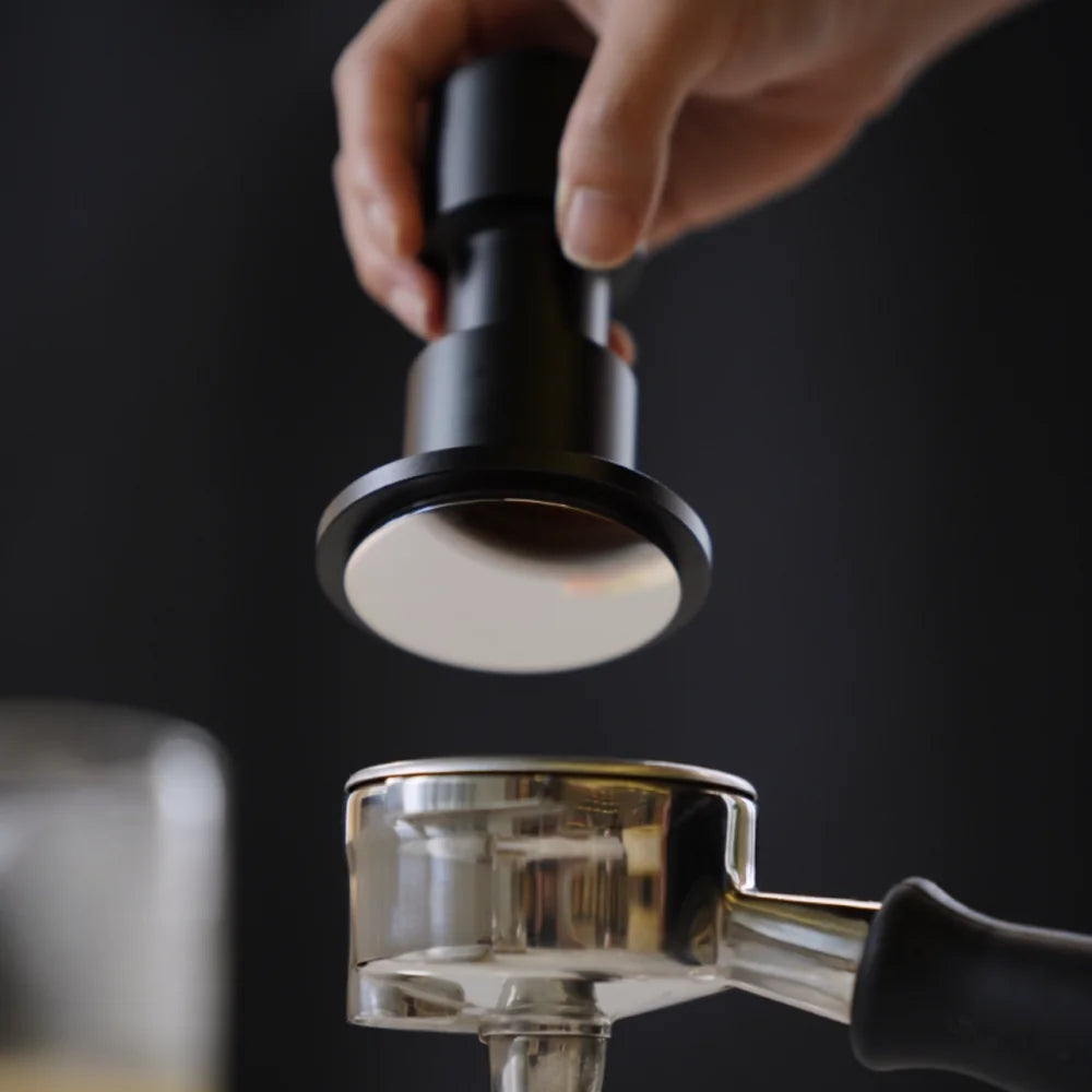 A hand lowering the black Impulse Impact Tamper with its polished stainless steel base over a portafilter against a dark background